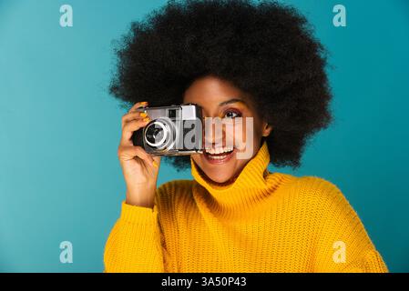 Femme noire souriante avec un afro prend une photo sur un fond de studio bleu. Un portrait vibrant adapté aux campagnes de style de vie, de mode et de photographie. Banque D'Images