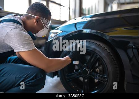 Mécanicien automobile afro-américain pulvérisant la surface de roue avec l'éclat de pneu accroupissant dans l'atelier de réparation automobile Banque D'Images