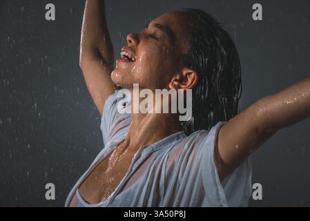 Femme asiatique debout avec les bras tendus et les yeux fermés profitant de la pluie. Un moment de style de vie agréable idéal pour le bien-être, les voyages et les campagnes en plein air. Idéal pour les campagnes sur la joie, la liberté et les humeurs rafraîchissantes. Banque D'Images