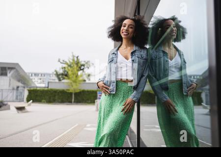 Joyeuse jeune femme afro-américaine aux cheveux bouclés, vêtue d'une veste en denim et d'une jupe verte à pois, sourit à la caméra en s'appuyant contre un mur. Un portrait de rue dynamique qui met en valeur la confiance, la mode et la vie urbaine. Idéal pour le style de vie, la mode et les accessoires. Banque D'Images
