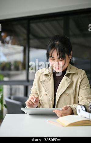 Asiatique femme freelance se concentre sur une tablette numérique dans un café confortable. Elle incarne le mode de vie du travail à distance avec une ambiance moderne et productive dans l'espace de travail. Idéal pour les sujets sur le freelancing, les nomades numériques et les arrangements de travail flexibles. Banque D'Images