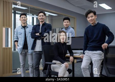 Un professionnel DE L'INFORMATIQUE chinois est assis à un bureau tandis que ses collègues se tiennent à proximité dans un bureau. Une scène d'équipe technologique collaborative adaptée aux projets INFORMATIQUES, au développement de logiciels et aux partenariats d'affaires. Banque D'Images