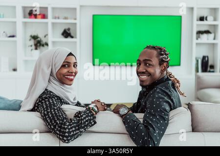 Couple africain se relaxant sur un canapé regardant la télévision avec un fond d'écran vert chroma. La scène comprend une femme portant un hijab, reflétant la diversité de la vie familiale. Banque D'Images