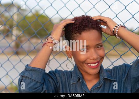 Femme colombienne souriante dans des vêtements décontractés profitant du temps dans un parc près d'une clôture en filet, capturant un style de vie en plein air détendu. L'espace de copie et une ambiance joyeuse conviennent à la marque de style de vie. Banque D'Images