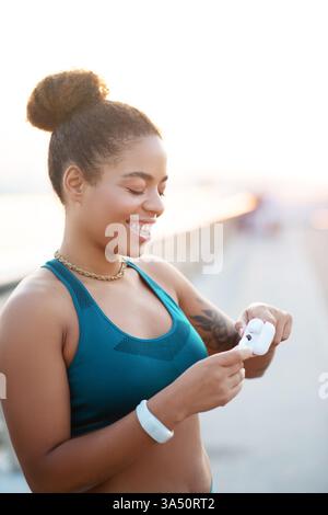 Femme excitée à la peau foncée sourit avant une séance d'entraînement, clairement énergisée pour l'exercice. Elle porte un équipement sportif et une smartwatch, incarnant la vie active. Idéal pour les campagnes de fitness, le style de vie en salle de sport et les images de santé motivantes. Banque D'Images