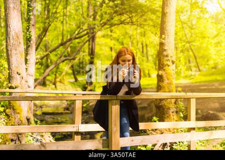 Femme photographe en automne, vêtue d'une veste noire, prenant des photos sur un pont en bois. Image saisonnière mettant en valeur la photographie, la nature et les paysages pittoresques du parc. Convient aux voyages, aux industries créatives et aux campagnes d'activités de plein air. Banque D'Images