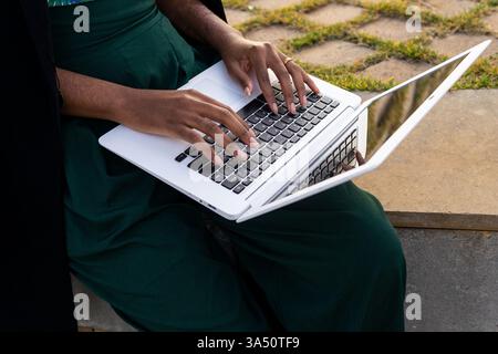 Photo en grand angle d'une femme afro-américaine élégante travaillant à distance depuis un parc, ordinateur portable ouvert sur son genou. Le recadrage et le réglage transmettent les vibrations nomades numériques, la mobilité et le travail extérieur contemporain. Banque D'Images