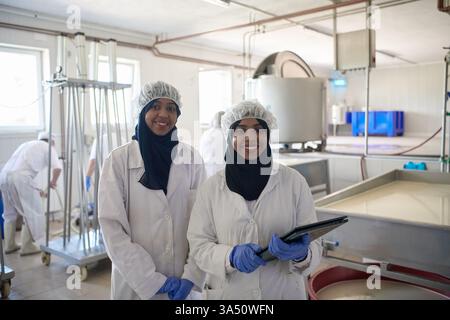 Des travailleuses du moyen-Orient souriantes en hijab et en uniformes blancs dans une usine de production laitière. Travail d'équipe et imagerie de fabrication moderne axée sur la production de fromage et de produits laitiers. Adapté aux visuels d'entreprise, industriels et de l'industrie alimentaire locale. Banque D'Images