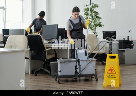 Vadrouille nettoyante féminine de race mixte dans un seau debout contre l'ordinateur de nettoyage de collègue mâle noir dans le bureau Banque D'Images