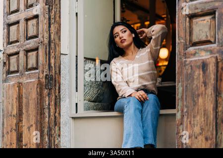 Jeune femme hispanique dans des vêtements décontractés touche ses cheveux tout en regardant loin près d'un rebord de fenêtre de café. Portrait de style de vie décontracté parfait pour la mode, la beauté ou les campagnes culturelles de café contemporain. Idéal pour le contenu des médias sociaux, le style de marque ou les blogs de style de vie en milieu urbain. Banque D'Images