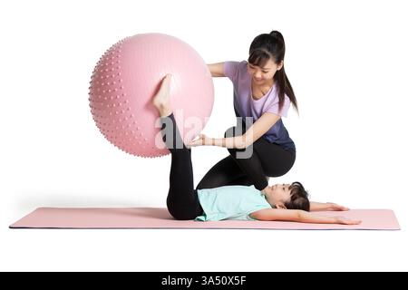 Mère et fille chinoises font de l'exercice ensemble dans un studio sur fond blanc. La scène joyeuse comprend un ballon de fitness et des conseils doux, démontrant la forme physique de la famille et un mode de vie sain. Idéal pour les activités parentales, le bien-être et les campagnes de fitness des enfants. Banque D'Images
