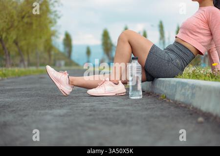 Vue latérale d'une athlète féminine reposant sur le trottoir après avoir couru, avec une bouteille d'eau en plastique à proximité. La pause capte le repos, la forme physique et un style de vie actif. Banque D'Images