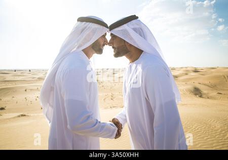 Les hommes arabes portant des kanduras se serrent la main dans un décor désertique pendant la journée. Le moment transmet amitié, tenue culturelle et collaboration dans un paysage moyen-oriental. Banque D'Images
