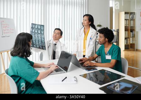 Équipe médicale examinant les radiographies du rachis pendant que les infirmières tapent sur les ordinateurs portables dans un environnement hospitalier. Parfait pour les soins de santé, les communications hospitalières et les visuels multidisciplinaires du travail d'équipe. Idéal pour les publications médicales, les concepts de soins aux patients et la marque clinique. Banque D'Images