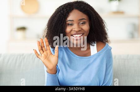 Portrait d'une femme afro-américaine souriante saluant tout en étant assise sur un canapé à la maison. La pose amicale et accueillante fonctionne bien pour les communications, le service à la clientèle et les contextes de style de vie. Idéal pour les intérieurs, les médias sociaux et les concepts d'accueil. Banque D'Images