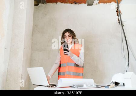 Architecte confiant Forewoman sur un chantier de construction utilise un smartphone tout en examinant les plans, portant un casque de sécurité. La scène exprime le leadership dans l'industrie du bâtiment et un environnement professionnel soucieux de la sécurité. Banque D'Images
