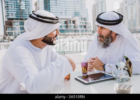 Souriants hommes d'affaires du moyen-Orient et du Caucase portant des kanduras assis dans un café en plein air partageant le thé. Ce moment d’affaires interculturel est adapté à la collaboration internationale et aux thèmes de l’hospitalité. Banque D'Images