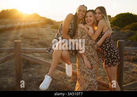 Une paire diversifiée d'amis partagent un câlin chaleureux sur une clôture en bois surplombant la côte au coucher du soleil. Le moment capture une amitié joyeuse et des vibrations estivales insouciantes le long du littoral. Parfait pour les campagnes de style de vie ou de voyage mettant en valeur la diversité, la joie et les moments côtiers. Banque D'Images
