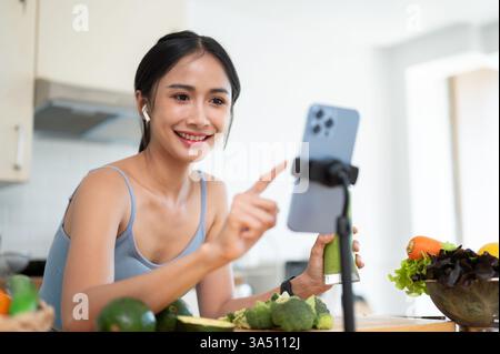 Une influenceuse asiatique dans les vêtements de gymnastique diffuse en direct depuis sa cuisine, partageant une recette de smoothie vert sain. La vidéo met en lumière la création de contenu sur les réseaux sociaux, la nutrition et un mode de vie sain et joyeux. Banque D'Images