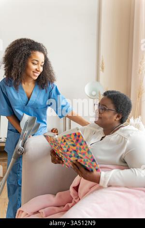 Deux femmes noires partagent un moment chaleureux de santé à la maison, avec une infirmière parlant à un patient sur un canapé. Cette scène met en lumière les soins à domicile, le professionnalisme infirmier et le bien-être des patients dans un cadre domestique. Banque D'Images