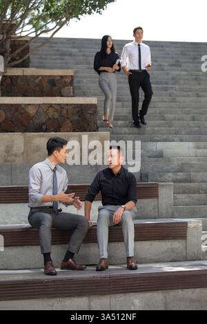 Deux hommes d'affaires chinois sont assis sur un banc de béton dans un parc de jour, engagés dans une discussion. La réunion en plein air transmet le leadership, le travail d'équipe et la collaboration professionnelle dans un cadre urbain. Convient pour les histoires sur les discussions d'affaires, le réseautage et les professionnels urbains. Banque D'Images
