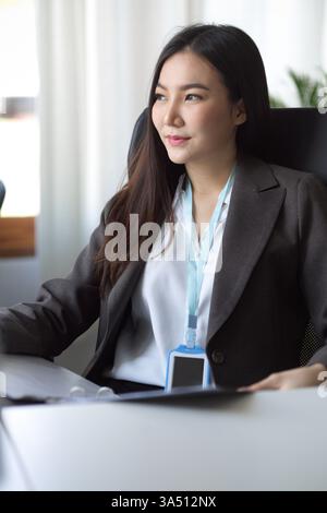 Femme d'affaires asiatique regardant loin tout en tenant un classeur à un bureau dans un bureau moderne. Un portrait poli et contemporain adapté aux thèmes de l'entreprise, du leadership ou du lieu de travail. Idéal pour les campagnes d'entreprise, le travail d'équipe et les services professionnels. Banque D'Images