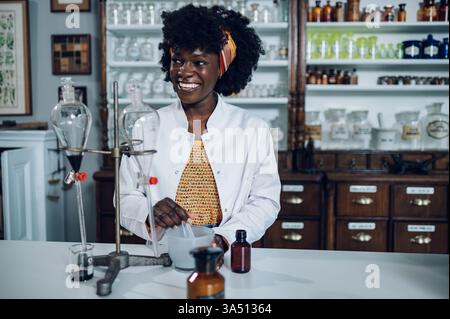 Joyeux chimiste afro-américain dans une apothicaire vintage mélange un composé dans un mortier et un pilon. Un portrait sur le thème de la santé et de la pharmacie. Banque D'Images