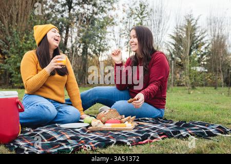 Groupe d'amis joyeux appréciant un pique-nique sur une prairie verte, partageant fromage, pain et boissons. Un moment de style de vie en plein air décontracté qui convient aux loisirs du week-end et aux thèmes de la restauration sociale. Idéal pour les campagnes sur les rassemblements en plein air, l'amitié et la vie estivale détendue. Banque D'Images