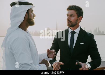 Deux hommes d'affaires, un moyen-oriental et un Caucasien, parlent près de la mer au coucher du soleil. Cette scène professionnelle avec une poignée de main et une discussion convient aux contextes corporatifs, immobiliers ou d'investissement. Banque D'Images