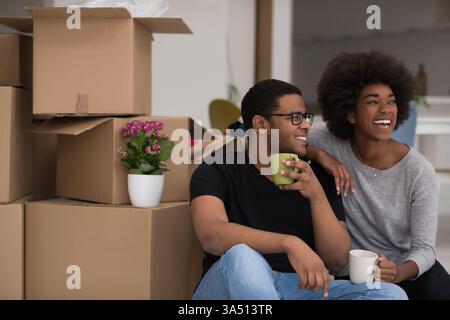 Un joyeux couple afro-américain se détend dans une nouvelle maison, assis sur le sol avec du café tandis que des boîtes de carton les entourent. La scène capture le déménagement, la vie à la maison et le bonheur. Banque D'Images