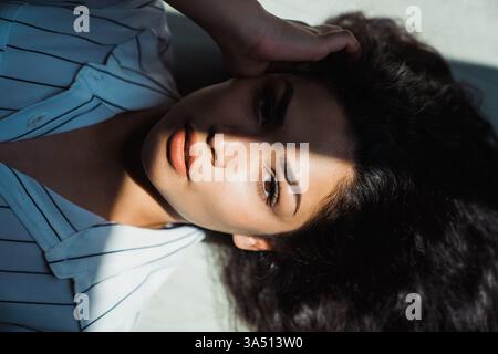 Femme hispanique sérieuse allongée sur un sol ensoleillé à la maison, main courant à travers ses cheveux noirs. Ce portrait utilise la lumière naturelle pour créer une image de style de vie calme et intime. Banque D'Images