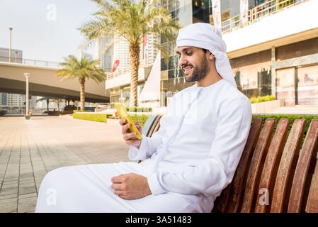 Jeune homme du moyen-Orient dans une kandura se promène à l'extérieur à Dubaï, mêlant tradition et vie urbaine moderne. Cette image de style de vie culturel convient aux voyages, aux affaires et aux caractéristiques culturelles régionales. Idéal pour les marchés du moyen-Orient et la narration de destinations. Banque D'Images