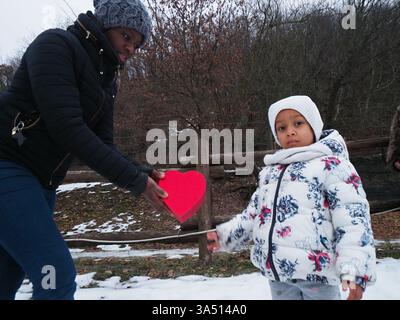Mère noire souriante donnant boîte en forme de coeur à sa fille avec les yeux fermés debout sur le parc enneigé pendant l'hiver Banque D'Images