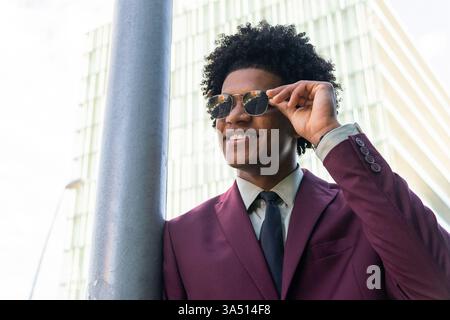 Homme d'affaires hispanique souriant avec les cheveux bouclés se penche sur un poteau de rue, touchant ses lunettes de soleil alors qu'il regarde au loin. Ce portrait urbain communique confiance, accessibilité et professionnalisme moderne. Banque D'Images