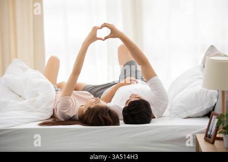 Couple chinois couché sur un lit dans une chambre à coucher faisant une forme de coeur avec leurs mains. Ce moment intime à la maison capture le romantisme, la convivialité et les vibrations détendues du week-end. Convient pour les rencontres, les relations ou les caractéristiques de la vie à la maison. Banque D'Images