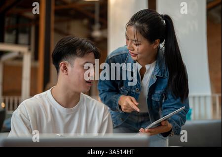 Deux jeunes collègues asiatiques dans un café discutent d’un projet et partagent des idées tout en travaillant ensemble. Ils sont assis à une table avec un ordinateur portable, mettant en valeur la collaboration et l'esprit d'entreprise. Idéal pour le coworking, l'étude et les thèmes d'affaires de café. Banque D'Images