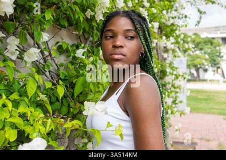 Femme afro-américaine confiante avec de longues tresses se tient à côté d'un mur couvert de plantes rampantes vertes, baignée de lumière estivale. Ce portrait extérieur convient à la mode, au style de vie et aux caractéristiques de style urbain. Banque D'Images