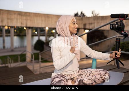 Femme arabe souriante portant des vagues de hijab tout en enregistrant une vidéo sur des escaliers en bois extérieurs. Cette image vibrante de style influenceur convient aux médias sociaux, aux vlogging ou aux campagnes beauté et style de vie. Banque D'Images