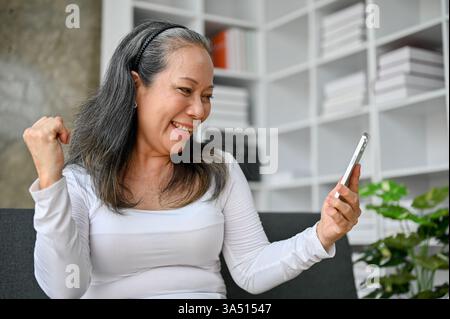 Une femme thaïlandaise âgée célèbre tout en regardant un écran de smartphone après avoir reçu de bonnes nouvelles à la maison. Ce moment chaleureux met en évidence l'utilisation de la technologie et le vieillissement joyeux dans la vie quotidienne. Banque D'Images