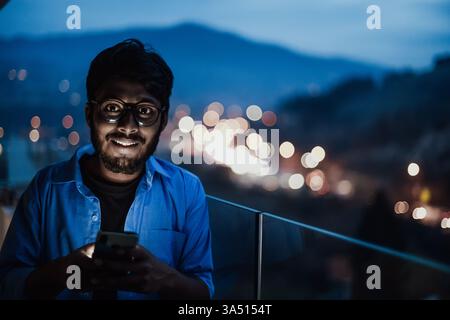 Homme indien avec des lunettes dans une chemise bleue utilise un téléphone portable la nuit sur un balcon donnant sur une rue de la ville. Cette scène technologique urbaine convient aux contextes télécom et télétravail. Idéal pour les applications, les services en ligne ou les images quotidiennes. Banque D'Images