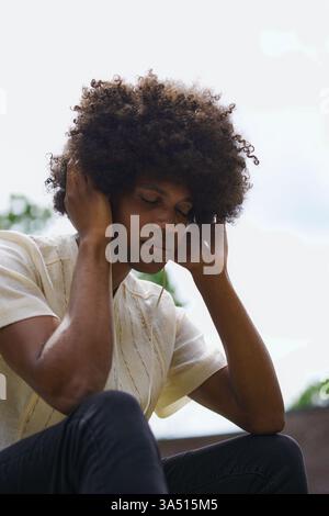 Détendu Black Man avec un Afro écoute de la musique sur des écouteurs dans un jardin extérieur par une journée ensoleillée. Il ferme les yeux, trempant dans l'instant présent. Parfait pour le bien-être, les loisirs et les visuels de style de vie en extérieur. Banque D'Images