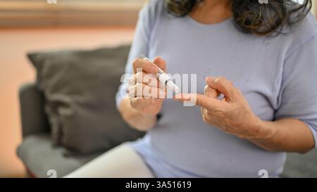 Une femme aînée asiatique vérifie son taux de sucre dans le sang avec un glucomètre lorsqu'elle est assise sur un canapé à la maison. Cette scène de soins de santé et de diabète met en évidence la surveillance de routine et les soins aux aînés dans un cadre de salon. Convient aux sujets médicaux, de bien-être et de santé à domicile. Banque D'Images