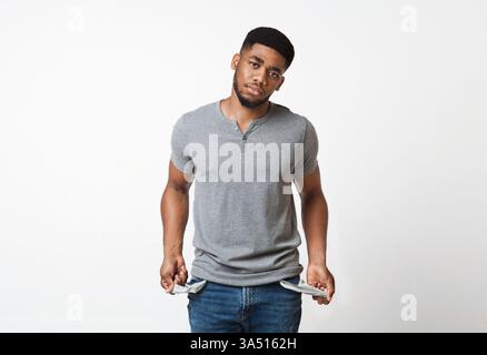 Homme afro-américain sans emploi debout dans un studio blanc, affichant des poches vides pour transmettre la pauvreté et le stress financier. Une image conceptuelle forte pour les discussions sur le chômage, la dette et les défis économiques. Convient pour les histoires éditoriales et les campagnes de sensibilisation sur les difficultés financières. Banque D'Images