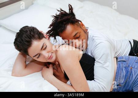 Un homme afro-américain embrasse sa petite amie caucasienne par derrière alors qu'il est allongé sur un lit dans une chambre. Un moment de vie chaleureux et intime qui convient à la romance et à la narration de la vie de couple. Banque D'Images