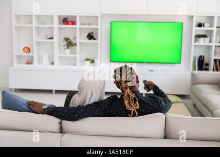 Couple africain se relaxant sur un canapé regardant la télévision ensemble, avec un fond d'écran vert chroma. La femme porte un hijab, reflétant un cadre familial diversifié. Idéal pour le style de vie familial, le divertissement à domicile et la publicité inclusive. Banque D'Images