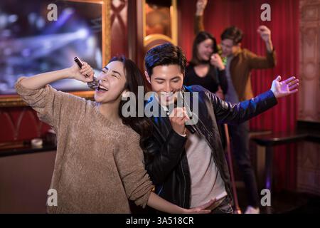Des amis chinois joyeux chantent au karaoké dans une discothèque sous les lumières de la discothèque. La scène intérieure montre un groupe de quatre personnes profitant d'une soirée animée, parfaite pour la vie nocturne et les thèmes musicaux. Utile pour le divertissement et les visuels du style de vie urbain asiatique. Banque D'Images