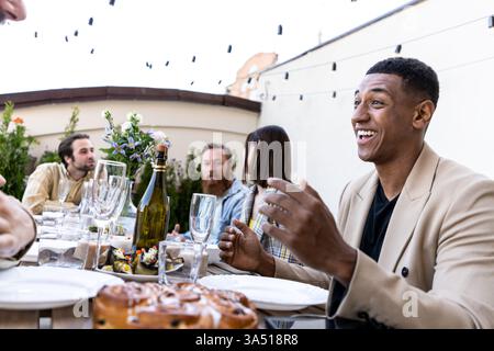 Un groupe multiethnique d'amis et de famille dîne sur une terrasse sur le toit lors d'une réunion à la maison. Les cordes lumineuses et l'atmosphère chaleureuse rendent cette image idéale pour le style de vie, la célébration et les campagnes de convivialité. Banque D'Images