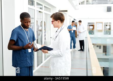 Vue de côté portrait d'une femme médecin en discussion avec un assistant dans un couloir de clinique moderne. Cette image de santé met en évidence le personnel médical professionnel, le travail d'équipe et un environnement clinique propre avec un espace de copie pour les légendes ou le texte. Idéal pour les sujets hospitaliers, cliniques et de soins de santé mettant en vedette divers professionnels. Banque D'Images