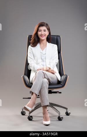 Femme d'affaires chinoise souriante assise jambes croisées sur une chaise de bureau dans un studio avec un fond gris. Un portrait professionnel adapté au leadership, aux communications d'entreprise et aux sujets d'autonomisation des femmes. Prise de vue studio propre avec espace de copie pour la marque. Banque D'Images