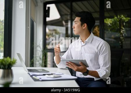 Homme d'affaires asiatique millénaire prospère regardant par la fenêtre tout en sirotant un café à son bureau. La pose réfléchie et le décor de bureau contemporain conviennent aux entreprises, aux dirigeants et aux campagnes d'entreprise. Banque D'Images